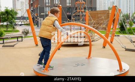 Junge dreht sich auf dem Karussell auf dem öffentlichen Spielplatz. Aktives Kind, Sport und Entwicklung, Kinder spielen im Freien. Stockfoto