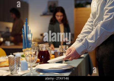 Nahaufnahme eines jungen Mannes, der Teller auf den Tisch stellte, der für das Hanukkah-Abendessen serviert wurde, während er seiner Familie bei der Vorbereitung half Stockfoto