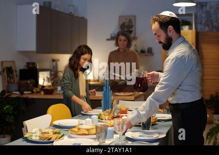 Seitenansicht eines bärtigen Mannes in weißem Hemd, der beim Servieren des Esstisch mit Tellern und einer Glaskanne mit hausgemachtem Saft für die Hanukkah-Feier hilft Stockfoto