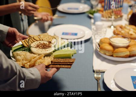 Konzentrieren Sie sich auf einen Teller mit frischer Gurke, Matzo, Pitabrot und Humus in einer kleinen Glasschale, die von einem jungen jüdischen Mann gehalten wird, der einen Tisch zum Abendessen serviert Stockfoto
