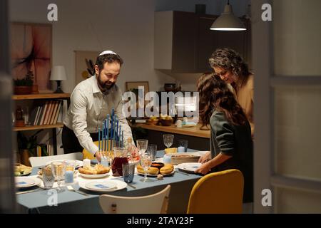 Jüdische dreiköpfige Familie serviert einen Tisch für das Hanukkah-Abendessen mit hausgemachten Speisen im Wohnzimmer, während sie sich auf den Empfang der Gäste vorbereitet Stockfoto