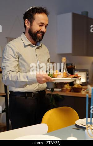 Ein lächelnder bärtiger Mann hält einen Teller mit einer Flasche Öl und hausgemachten Snacks, während er einen Tisch für das Hanukkah-Abendessen in der Küche serviert Stockfoto