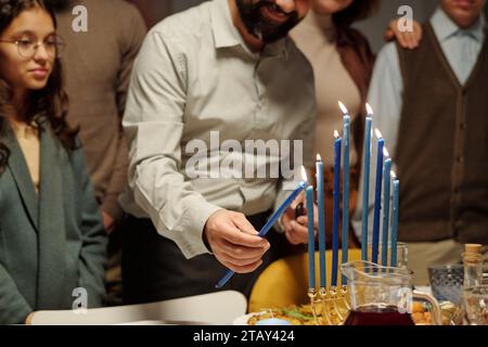 Ein bärtiger Mann zündet eine Kerze der Menora auf einer Hanukkah-Party an, während er neben seinem Tisch stand, der mit seiner Tochter und der anderen Familie serviert wurde Stockfoto