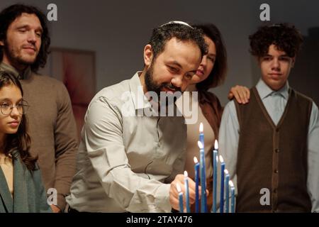 Reifer Mann in jüdischem Schädelkappen, der brennende blaue Kerze auf den Leuchter der Menora setzt, während er Hanukka zu Hause mit seiner Familie feiert Stockfoto