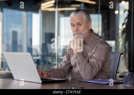 Porträt eines älteren grauhaarigen Geschäftsmannes, der im Büro am Tisch sitzt, an einem Laptop arbeitet und ernsthaft die Kamera betrachtet. Stockfoto