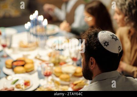 Seitenansicht des Kopfes eines reiferen bärtigen Mannes in Yarmulke mit Davidssternen, die vor der Kamera am Tisch sitzen und zum Hanukka-Abendessen serviert werden Stockfoto