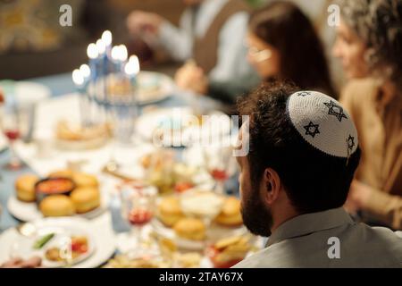 Seitenansicht des Kopfes eines reiferen bärtigen Mannes in Yarmulke mit Davidssternen, die vor der Kamera am Tisch sitzen und zum Hanukka-Abendessen serviert werden Stockfoto