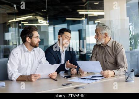 Drei verschiedene Geschäftsleute, die sich gemeinsam an einem Besprechungstisch im Büro unterhalten, Geschäftsleute in Business-Anzügen, die mit dem Chef des Geschäftsinhabers über finanzielle Indikatoren und Erfolge diskutieren. Stockfoto