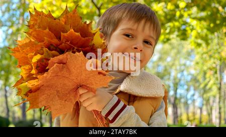 Porträt eines fröhlich lächelnden Jungen, der sich hinter einem Haufen gelber Ahornblätter im Herbstpark versteckt. Kinder im Freien, glückliche Kinder und Kindheit, Herbstlandschaft Stockfoto