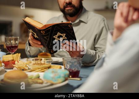 Abgeschnittener Schuss eines bärtigen Mannes mit offener Tora, der Psalmen oder andere Verse aus dem Alten Testament liest, während er am Tisch saß Stockfoto