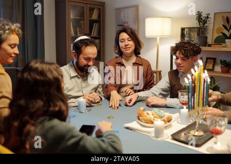 Junge Frau, die Münze auf den Tisch schiebt, während sie ein Freizeitspiel spielt, während Mitglieder ihrer Familie am Tisch mit einem Menorahenkerzenhalter sitzen Stockfoto