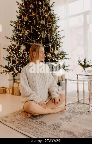 Blonde schwangere Frau, die sanft ihren Bauch vor dem Hintergrund des weihnachtsbaums berührt Stockfoto