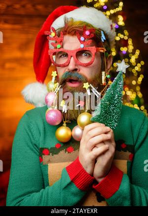 Bärtiger Mann mit Weihnachtsmütze mit dekorativen Weihnachtskugeln im Bart. Überraschter Kerl in Neujahrskleidung und Partybrille mit kleinem Weihnachtsbaum Stockfoto