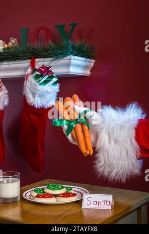 Der Weihnachtsmann hält Karotten für das Rentier neben Milch und Keksen mit Weihnachtsstrümpfen, die an einem weißen Kamin hängen Stockfoto