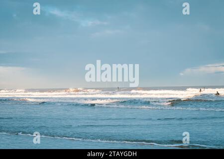 Beobachten Sie die weißen Wellen am Longsands Beach Tynemouth mit Surfern, die an einem eisigen Wintertag mit Schnee am Strand und hellblauem Himmel Wellen fangen. Stockfoto