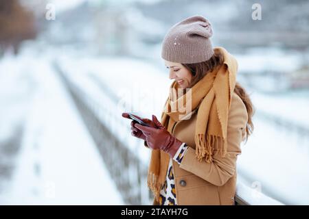 Fröhliche, moderne 40-jährige Frau mit braunem Hut und Schal in Kamelmantel mit Handschuhen, die im Winter draußen in der Stadt mit dem Smartphone Textnachrichten sendet. Stockfoto