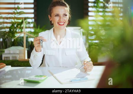 Umweltfreundliches Immobiliengeschäft. Glückliche elegante 40-jährige Maklerin in modernem grünen Büro in weißer Bluse mit Taschenrechner, Zwischenablage, Dokument und Schlüssel Stockfoto