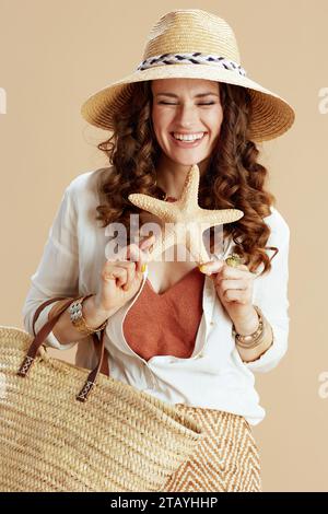Strandurlaub. Lächelnde elegante Hausfrau mittleren Alters in weißer Bluse und Shorts isoliert auf beigefarbenem Hintergrund mit Seestern, Strohbeutel und Strohhut. Stockfoto