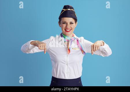 Lächelnde, moderne weibliche Flugbegleiterin vor blauem Hintergrund in Uniform, die auf den Kopierraum zeigt. Stockfoto