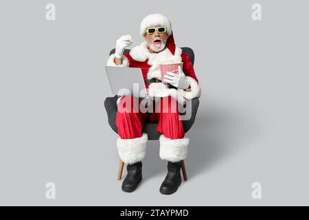 Weihnachtsmann in 3D-Brille mit Eimer mit leckerem Popcorn und Laptop, der Film auf grauem Hintergrund ansieht Stockfoto