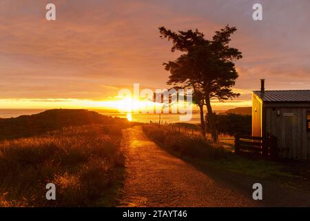 Orangefarbener Sonnenaufgang auf der Isle of Skye über einer Straße mit einem Baum und einem Haus in Silhouette Stockfoto