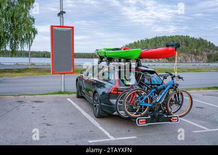 Schwarzes persönliches Auto mit zwei montierten Mountainbikes auf der Rückseite und zwei Kajaks auf der Oberseite. Grüne und rote Kajaks. Das Auto steht auf einem leeren Parkschild Stockfoto