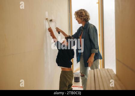 Heimwerkerarbeit zu Hause mit einer kaukasischen Mutter und ihrem jungen Sohn, die zusammen eine Innenwand malen. Mutter und Kind, die eine Hausrenovierung durchführen und das Stockfoto