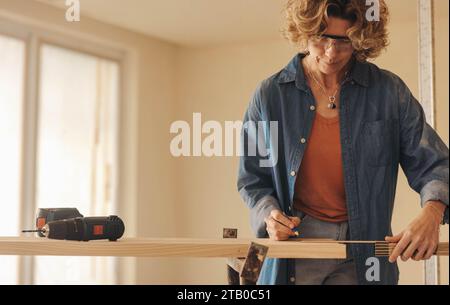 Reife weibliche Umbauerin, die an einem Küchen-Renovierungsprojekt arbeitet. Sie misst und markiert die Fußleisten mit einem Maßband und Bleistift. Karpfenqualifikation Stockfoto