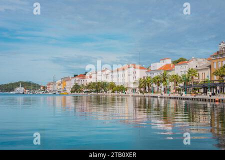 Promenade mit Palmen und anderen Bäumen und Häusern im Hintergrund in der Stadt Mali Losinj auf der kroatischen Insel Losinj an einem sonnigen Herbsttag. Stockfoto