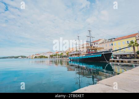 Promenade mit verankertem Segelboot, Palmen und anderen Bäumen und Häusern im Hintergrund in der Stadt Mali Losinj, auf der kroatischen Insel Losinj, auf einer sonne Stockfoto