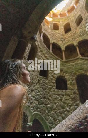 Frau, die von den majestätischen Steinbögen der Quinta da Regaleira in Sintra, Portugal, fasziniert aufblickt, von oben im Sonnenlicht getaucht, Stockfoto