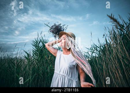 Mädchen mit hellen Zöpfen im Strohhut, der Blumenstrauß hält, Wind in der Natur Stockfoto