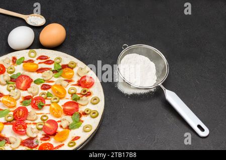 Runder Teigboden für Pizza. Lebensmittelzutaten zum Kochen von Pizza. Mehl im Sieb. Weizenzweig. Flache Lagen. Schwarzer Hintergrund. Stockfoto