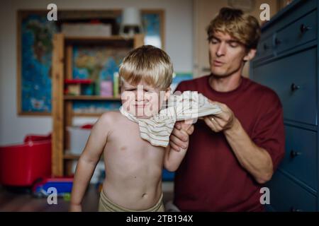 Vater zieht Hemd auf den kleinen Sohn und zieht sich morgens vom Schlafanzug. Der Junge ist traurig, weinend. Stockfoto