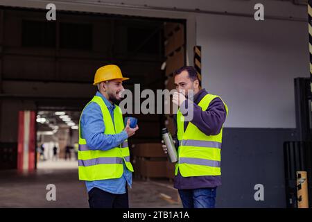 Porträt von zwei Arbeitern, die eine Pause von der Arbeit machen, Kaffee trinken, reden und sich ausruhen, auf der Laderampe stehen, im Freien. Kollegen trinken warmen Tee Stockfoto