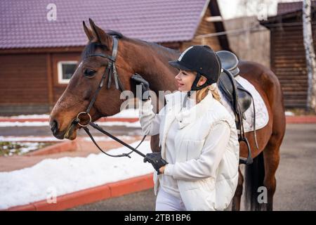 Lächelnde blonde Profi-Jockey, die neben dem Pferd steht. Freundschaft mit Pferd Stockfoto