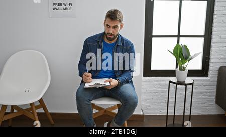 Der hübsche junge hispanische Mann, ein ernster Erwachsener mit Bart, entspannt sich in einem Stuhl, tief vertieft darin, einen Bericht auf einem Klemmbrett in einer ruhigen Inneneinrichtung zu schreiben Stockfoto