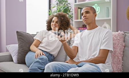 Ein wunderschönes Paar sieht sich fröhlich ihren Lieblingsfilm an, sitzt zu Hause auf der Couch, teilt ein Lächeln und lacht Stockfoto