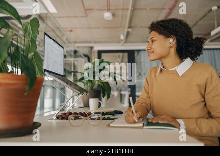 Lächelnde Architektin, die an einem Projekt arbeitet, während sie mit dem Laptop arbeitet und im Büro Notizen macht Stockfoto