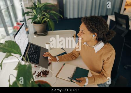 Lächelnde Geschäftsfrau, die an einem Projekt arbeitet, während sie im Büro einen Laptop benutzt und Notizen macht Stockfoto