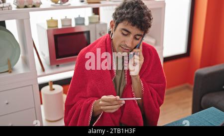 Junger hispanischer Mann, der auf einem Smartphone spricht und im Speisesaal ein Thermometer aussieht Stockfoto