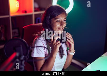 Junge schöne hispanische Frau streamer lächelnd zuversichtlich hält Kopfhörer im Spielsalon Stockfoto