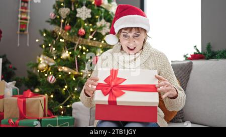 Reife hispanische Frau trägt weihnachtshut, die Geschenk mit überraschtem Gesicht zu Hause auspackt Stockfoto