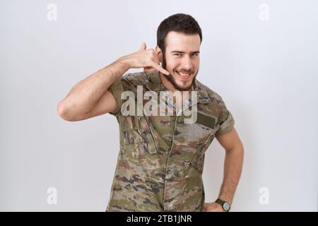 Junger lateinamerikanischer Mann in Tarnuniform, lächelnd, der Handygesten mit Hand und Fingern macht, als würde er am Telefon reden. Kommunizierende Konz Stockfoto