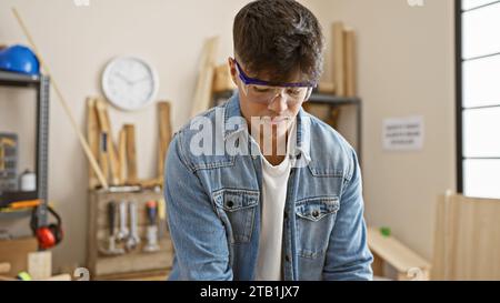 Hübscher, junger hispanischer Mann, der in einem Tischlerstudio groß steht – ein Porträt von Hingabe und Ernsthaftigkeit. Dieser mit Brille bekleidete, professionelle Tischler Stockfoto