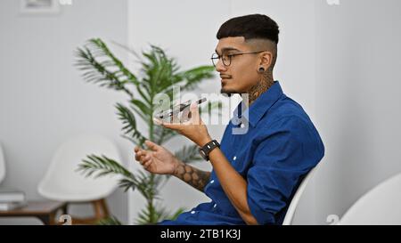 Junger lateinamerikanischer Mann, der in ernsthafte Gespräche vertieft ist, eine Sprachnachricht über sein Smartphone sendet und bequem im Wartezimmerstuhl sitzt Stockfoto