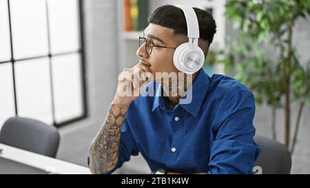Seriöser und stilvoller junger latin-Mann, der die Show im Büro leitet, Kopfhörer an, in Gedanken verloren, im Takt schwingt, während er die Geschäftswelt beherrscht Stockfoto