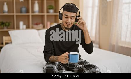 Selbstbewusster, tätowierter junger latin-Mann entspannte sich in seinem Bett, genoss seinen Morgenkaffee und vibrierte zur Musik in seinem gemütlichen Schlafzimmer Stockfoto