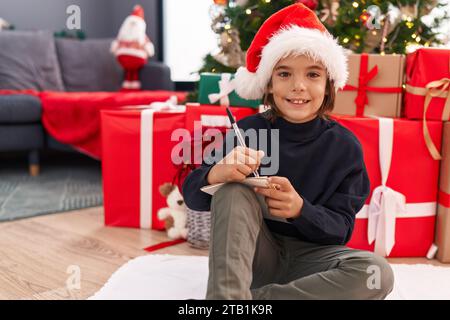 Entzückender hispanischer Junge, der den weihnachtsmann-Brief schreibt, der zu Hause am weihnachtsbaum sitzt Stockfoto