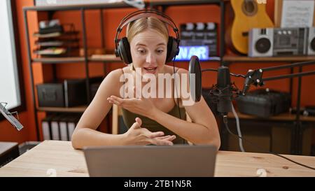 Junge blonde Radioreporterin, die im Podcast-Studio mit Kopfhörern und Laptop spricht Stockfoto